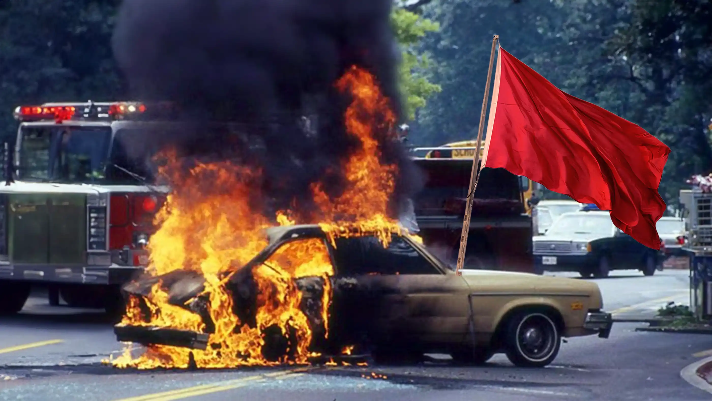 A Ford Pinto burns on the street with a firetruck in the background. A red flag sticks up from behind the burning vehicle.