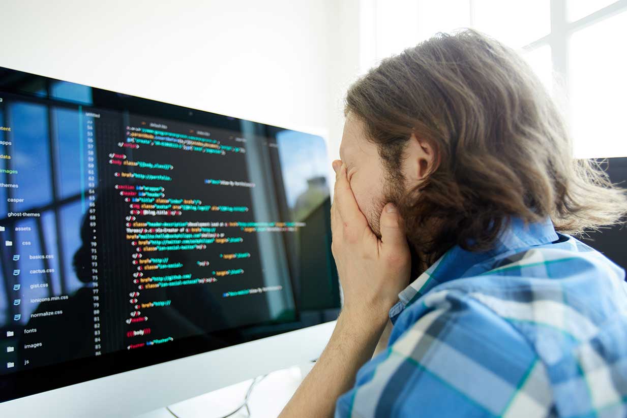 A frustrated developer burying his face in his hands in front of his computer screen filled with lines of code.