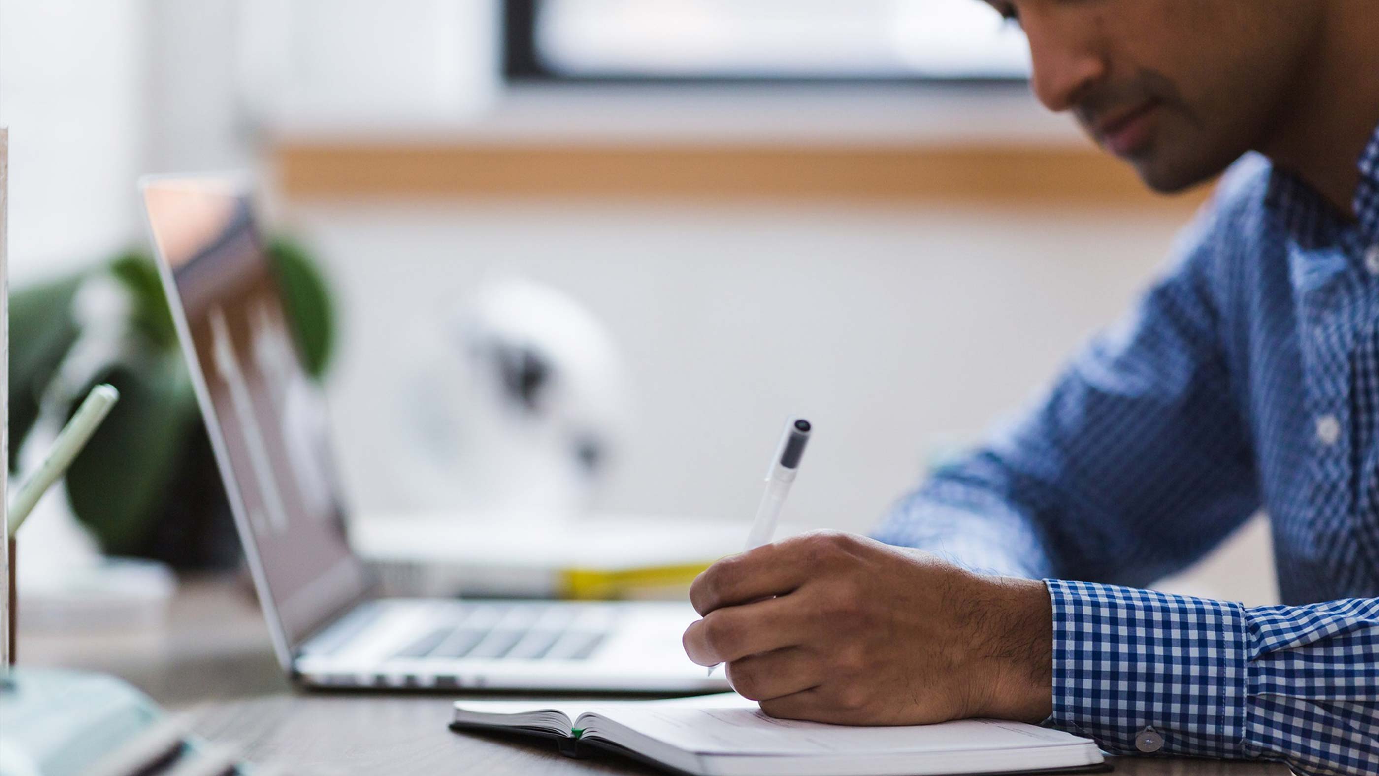 A man takes notes at his desk while taking an online educational course from his laptop. 