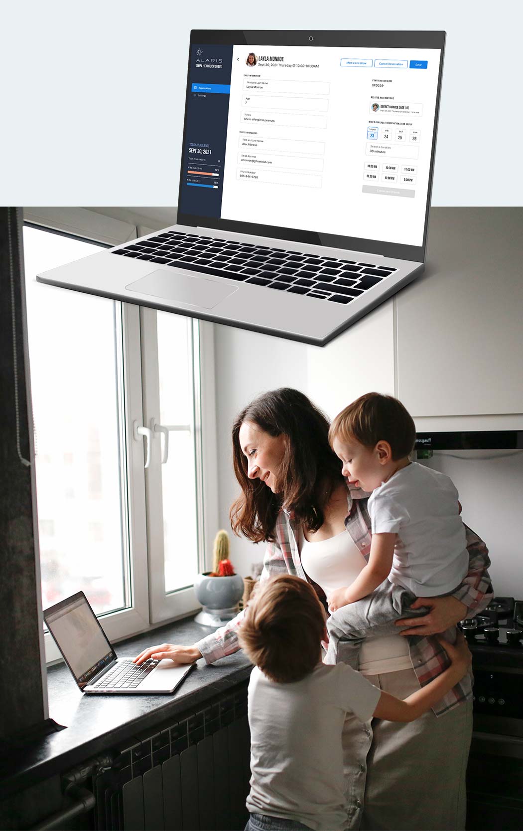 Woman holding children in her kitchen, booking an appointment on her laptop.