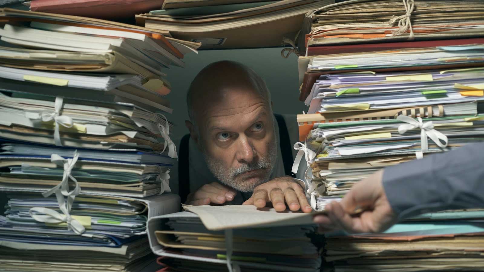 The old man hides in his fort made of paperwork and accepts more paperwork from someone through a window like a troll accepting a bridge toll.