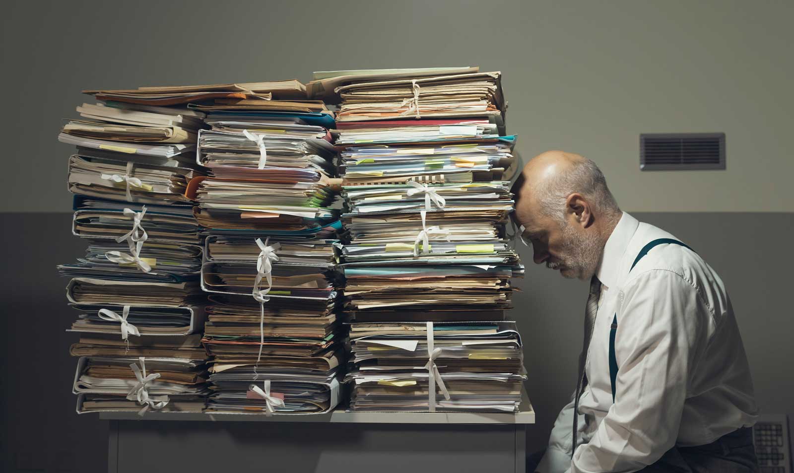 A man is resting his head against a dramatic mound of paperwork, frustrated and defeated.