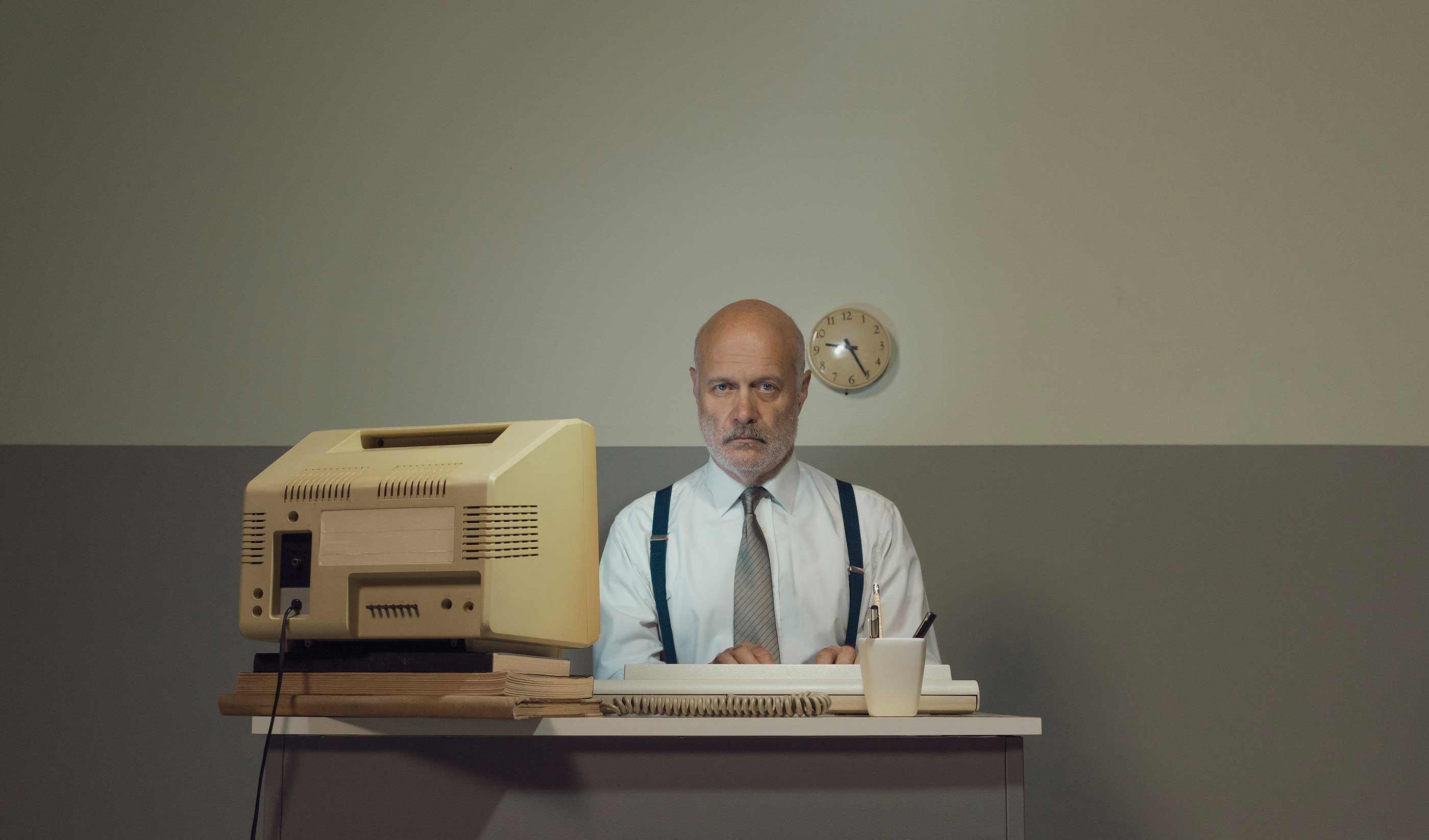 A clearly old man, both physically and fashionably, sits at an old desk with an old computer feeling old and sad.