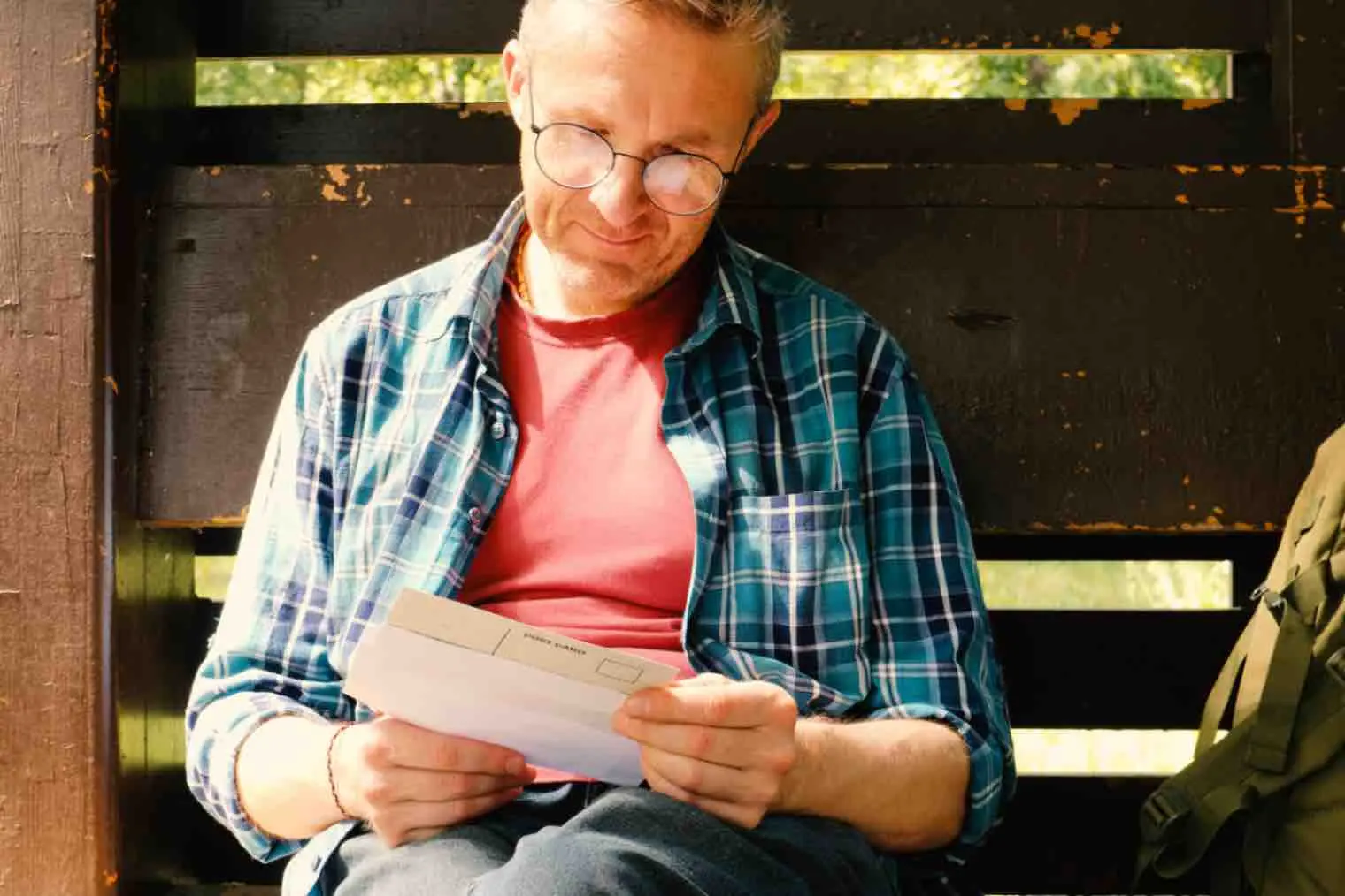 A grandfather is sitting on a bench smiling as he opens mail containing a postcard 