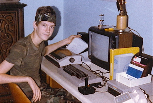 An image that appears to be from the 80s, featuring a teenage boy sitting next to a brick of a computer. 