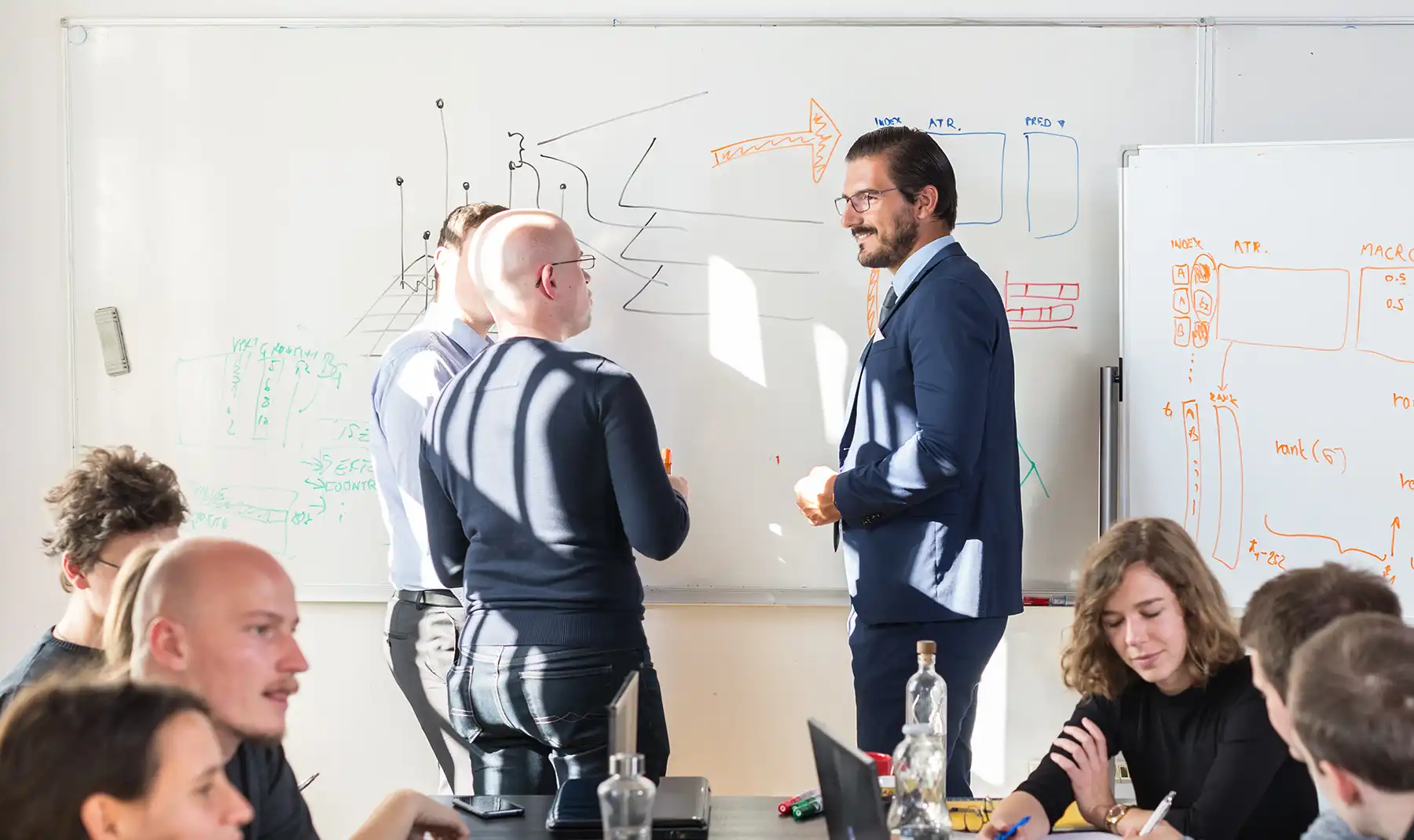 Three men in suits stand at a whiteboard, one of them smiles at the other two.