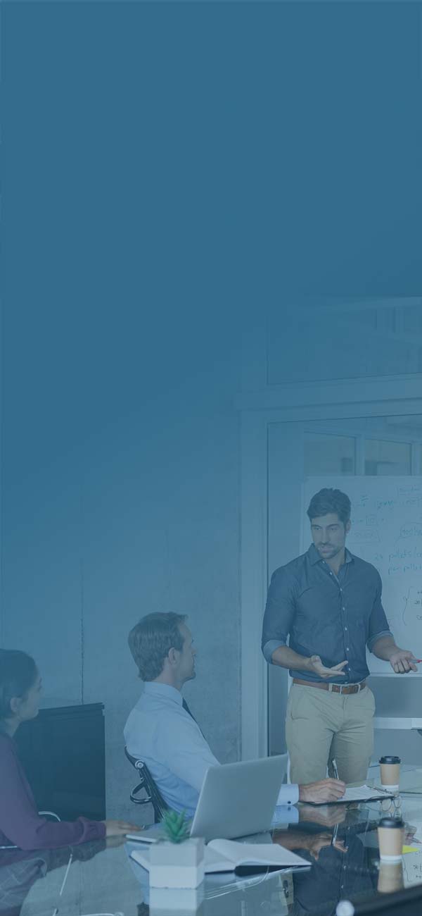A team of 5 people in a room with one man standing in front of a whiteboard, facilitating the group conversation.