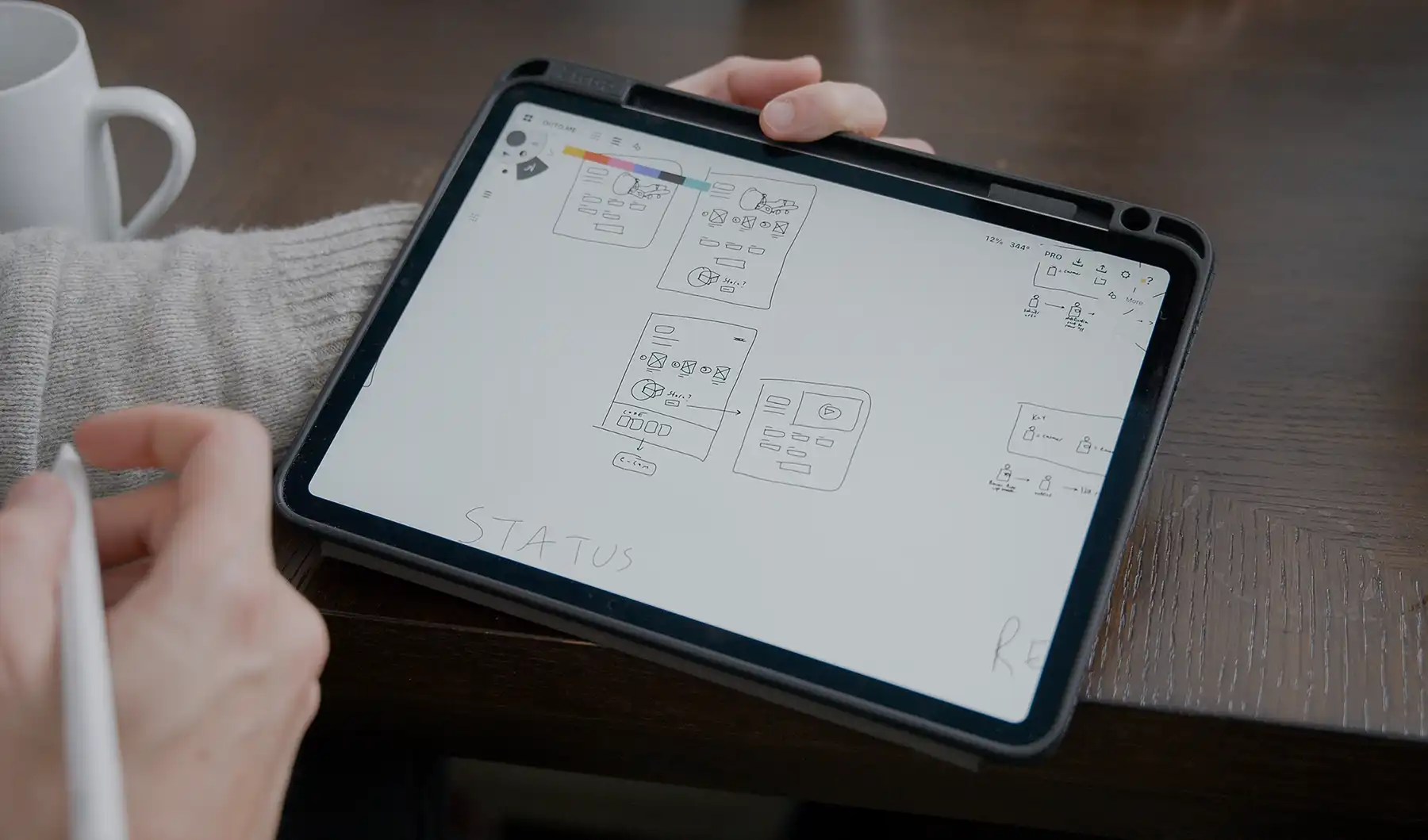 Wireframes are shown on the screen of a lead UX designer's tablet. He holds a stylus in his right hand. The word "status" is shown at the bottom of the screen.